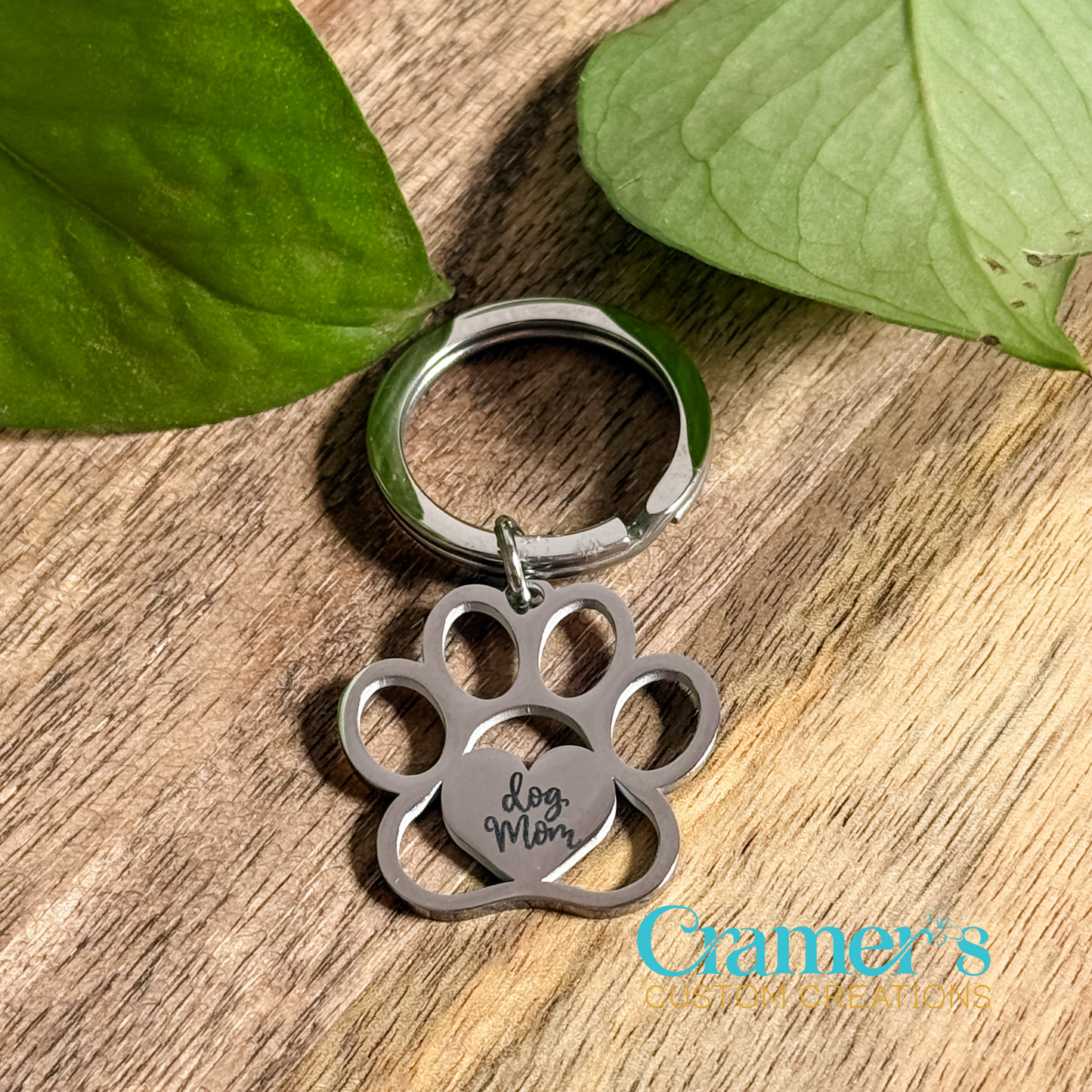 A paw print keychain with the inscription 'Dog Mom' on it, placed on a wooden surface with green leaves in the background.