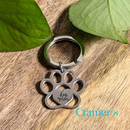 A paw print keychain with the inscription 'Dog Mom' on it, placed on a wooden surface with green leaves in the background.