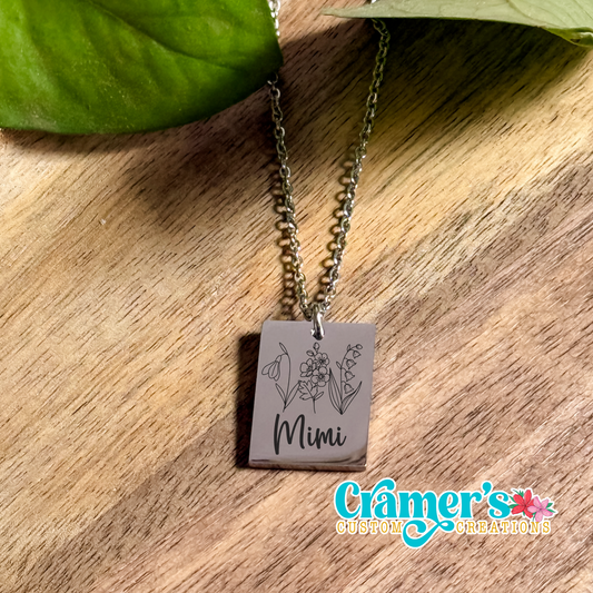 A stainless steel horizontal bar necklace with a pendant featuring a floral design and the name 'Mimi' engraved on it. The necklace is displayed on a wooden surface with a leafy background.