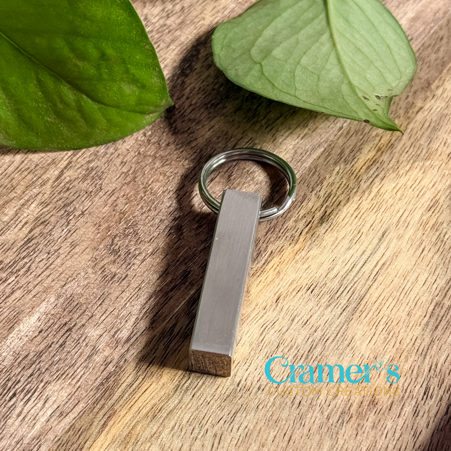 A stainless steel vertical bar keychain with a brushed finish on a wooden surface, next to green leaves.