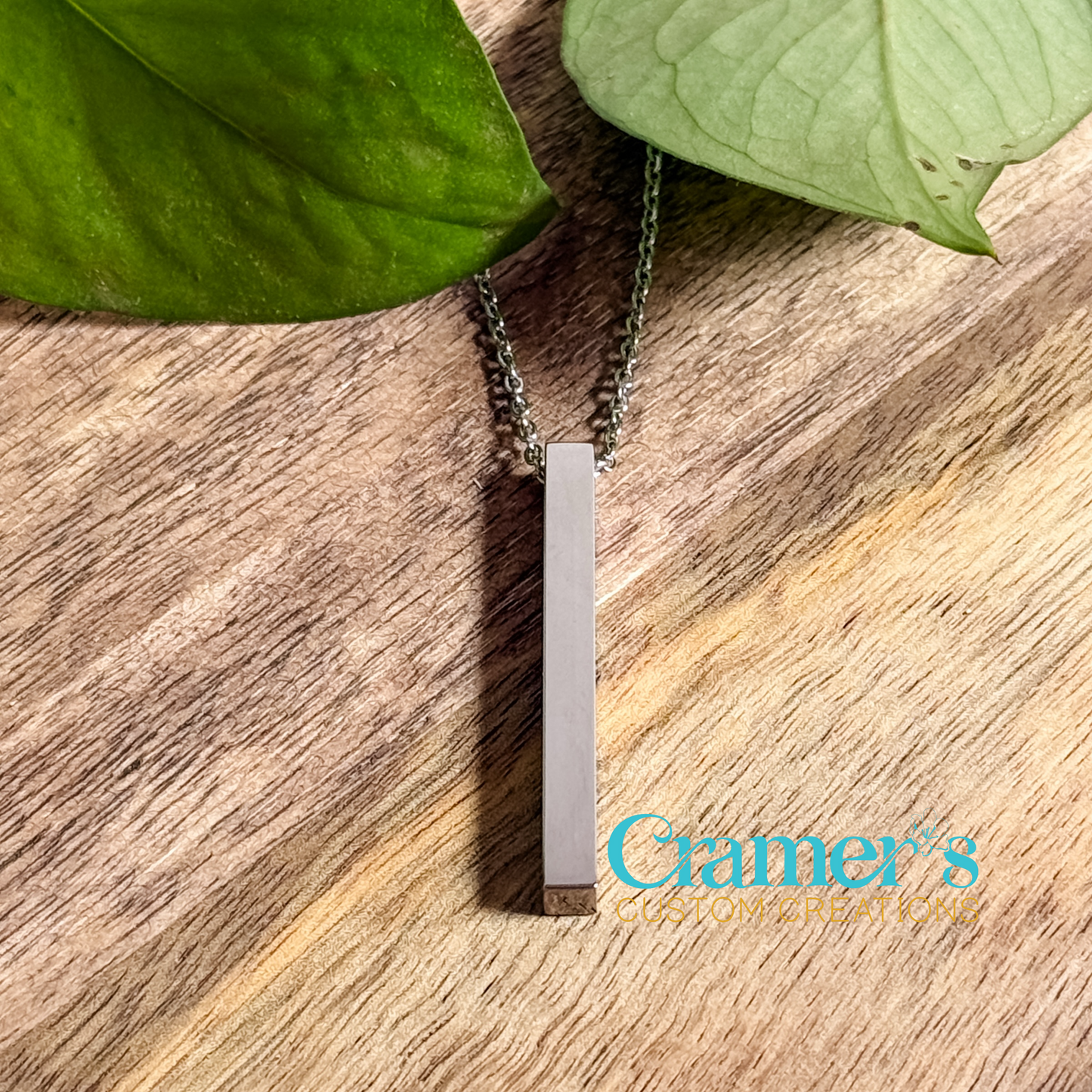 A vertical bar pendant necklace in silver color, with the chain displayed on a wooden surface surrounded by green leaves.