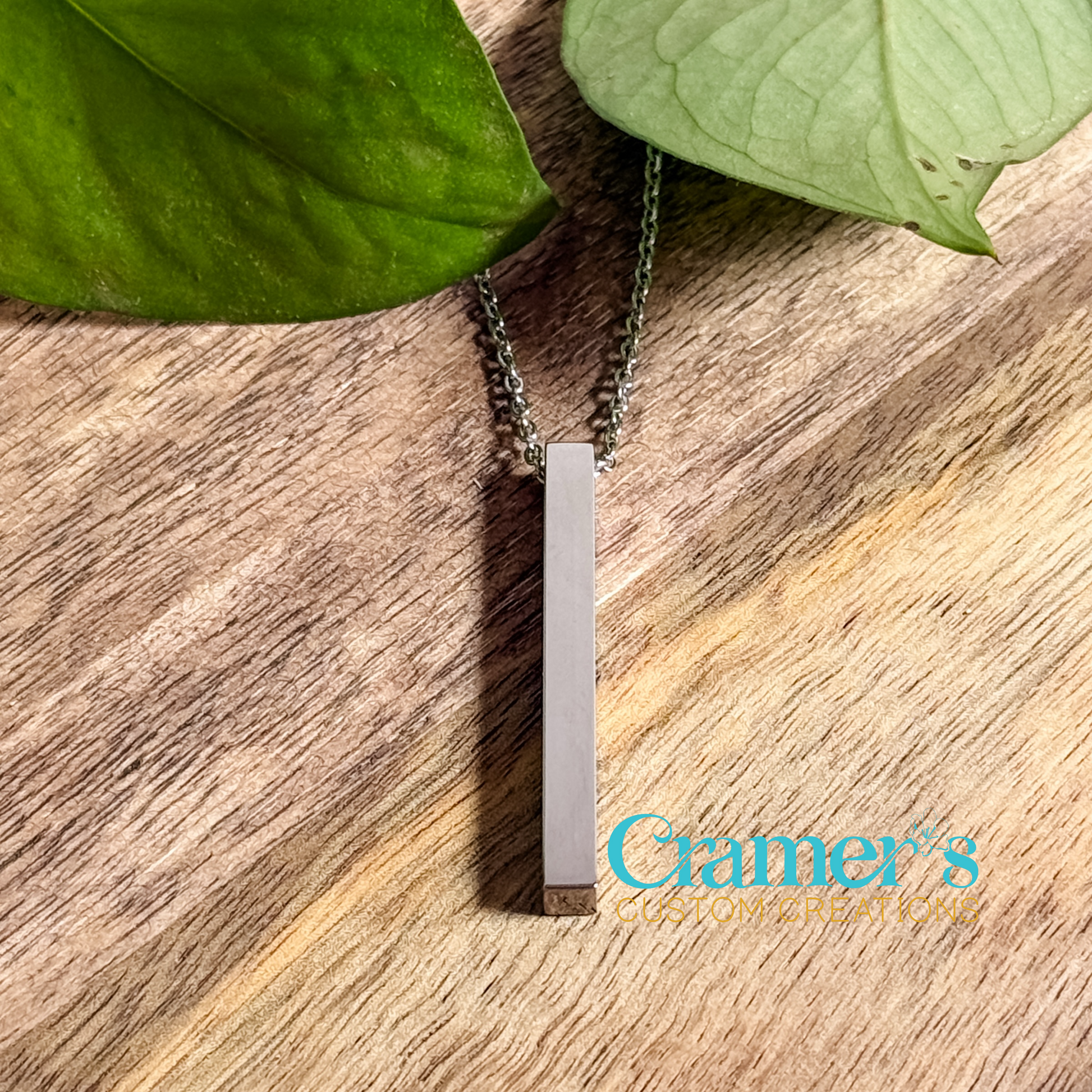 A vertical bar pendant necklace in silver color, with the chain displayed on a wooden surface surrounded by green leaves.