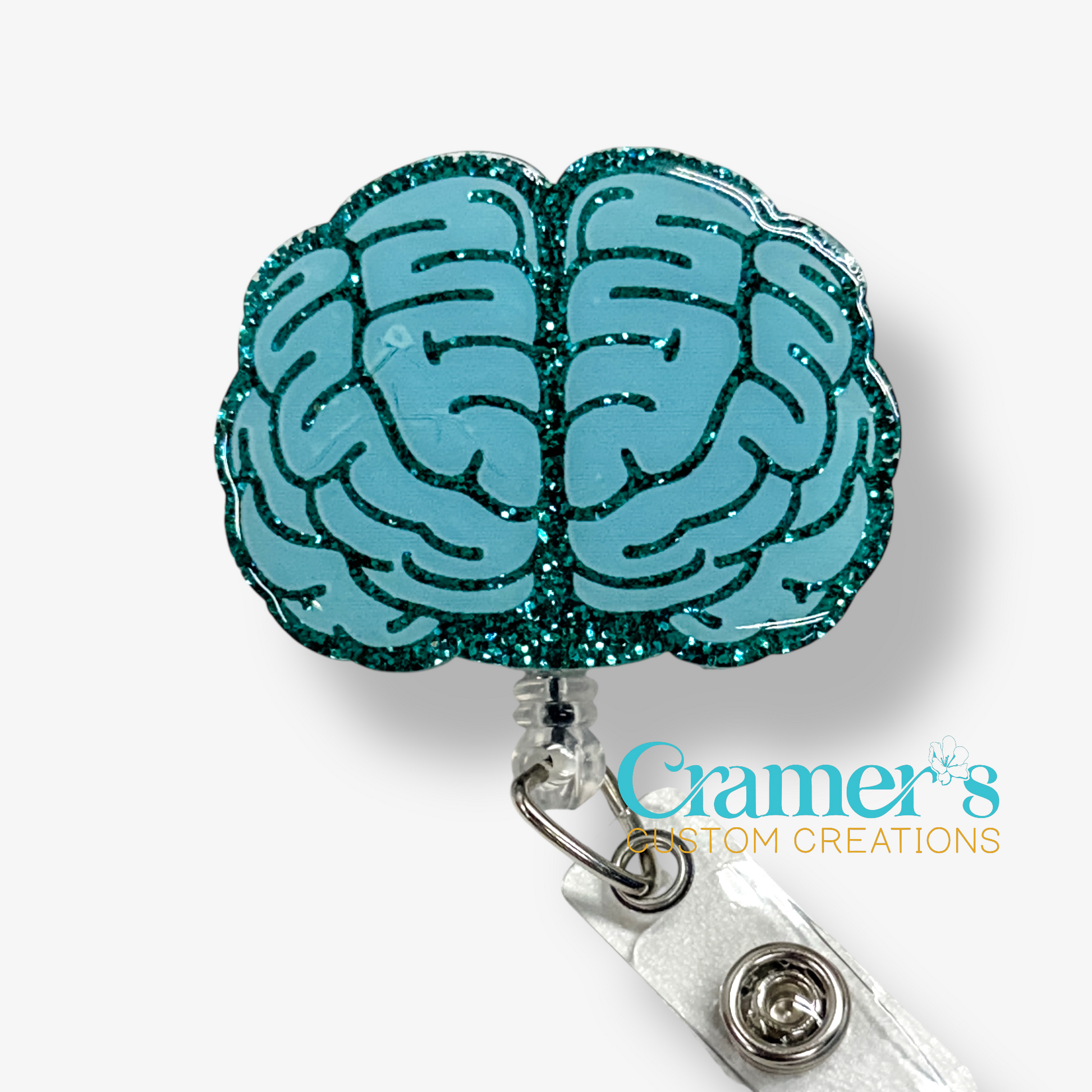 Brain-shaped badge holder with teal glitter design on a white background, featuring 'Cramer's Custom Creations' branding.