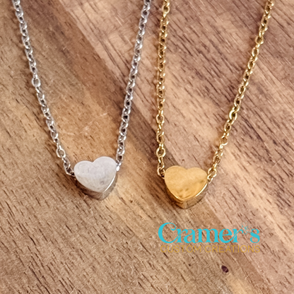 Two heart-shaped pendants on chains, one silver and one gold, on a wooden surface with 'Cramer's Custom Creations' branding.