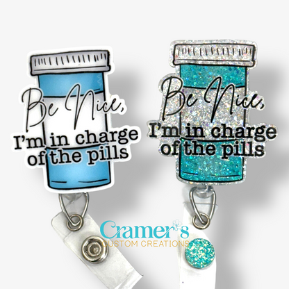 2 badge reels showing a glitter and a non glitter option of a badge with a pill bottle with the words be nice, i'm in charge of the pills