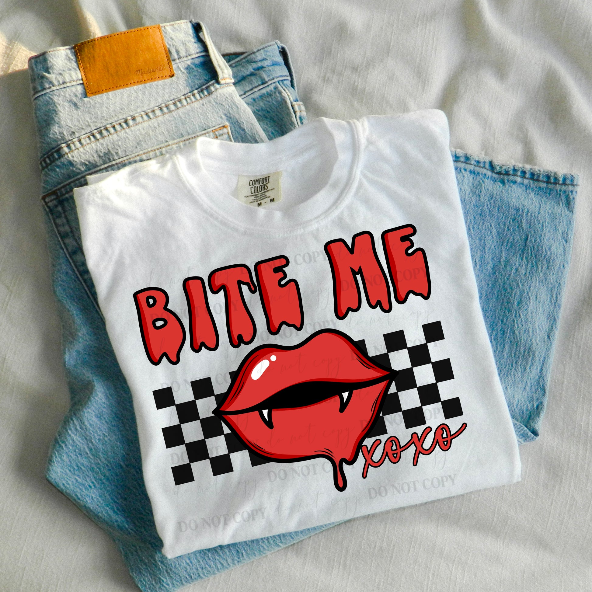 a white shirt with bit me on it with vampire lips on a checkered background.  shirt is laying on a pair of folded up jeans