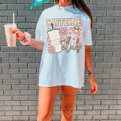 woman standing against a wall holding an iced coffee with a shirt that says caffeine club with a retro design