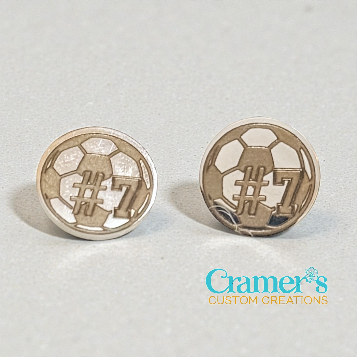 Pair of silver soccer ball earrings with '#7' on a light gray background, branded 'Cramer's Custom Creations'.