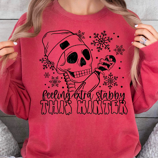 A pink t-shirt with a black and white skeleton design, surrounded by snowflakes, and the text 'Feeling Extra Stabby this Winter'.