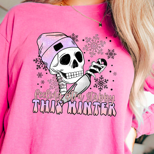 woman wearing a pink shirt that has a skeleton head wearing a beanie holding a knife that says feeling extra stabby this winter