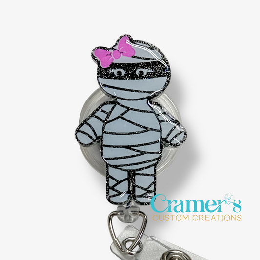 Mummy-shaped badge holder with a pink bow on a white background, featuring the brand 'Cramer's Custom Creations'.