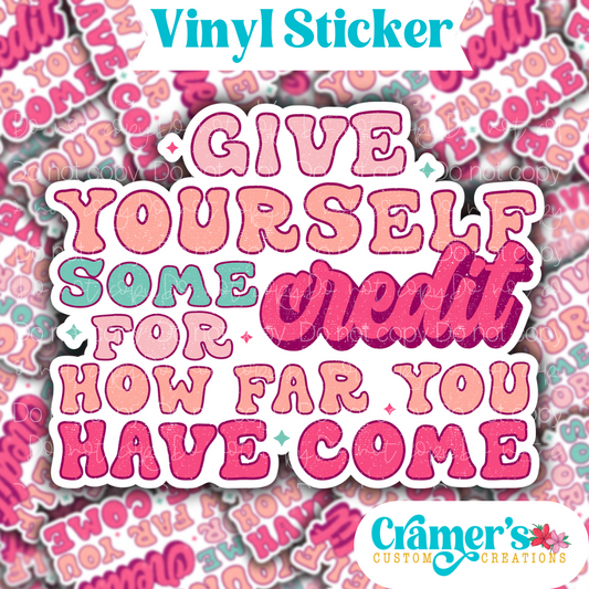 Sticker laying flat that says give yourself some credit for how far you have come on a background of the same sticker stacked up