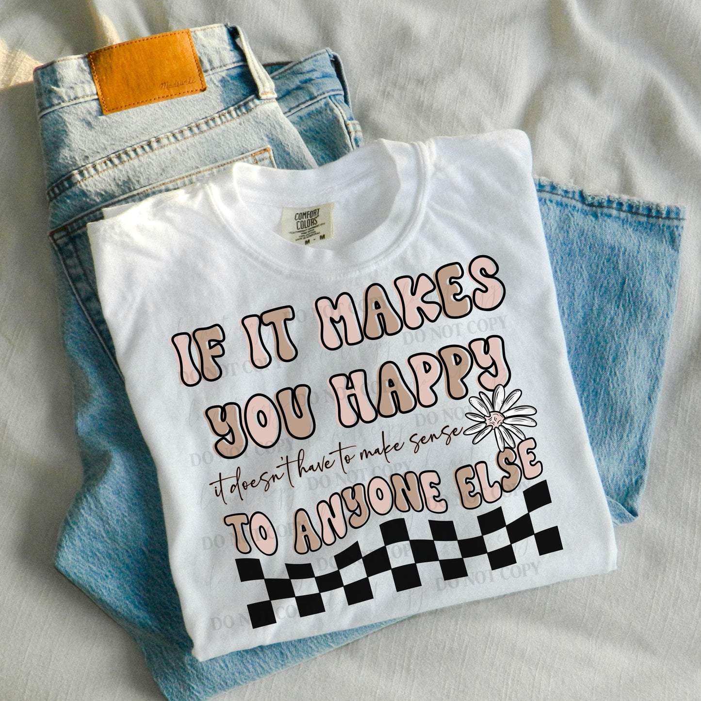 A white t-shirt with pink and black text and floral design, placed on a surface with blue jeans and a brown belt in the background.
