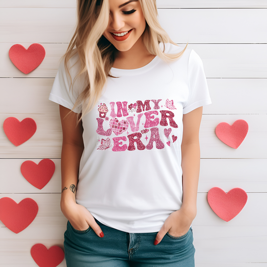 A person wearing a white t-shirt with a pink and red heart graphic and the text 'IN MY LOVER'S ERA'.