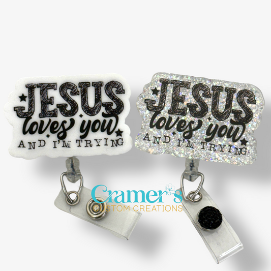 Two badge reels with acrylic coating and high quality glitter, one with a silver clip and the other with a black clip, both with the text 'JESUS LOVES YOU AND I'M TRYING' written in a bold, black font, surrounded by sparkle accents.