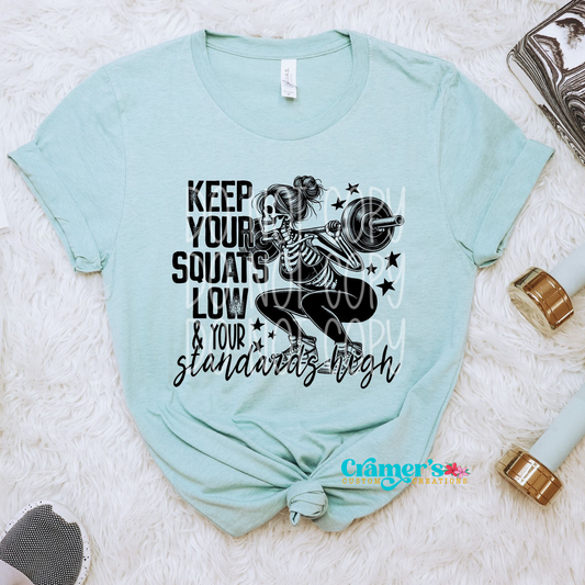 a blue shirt flat lay with a skeleton doing a squat saying keep your squats low and your standards high
