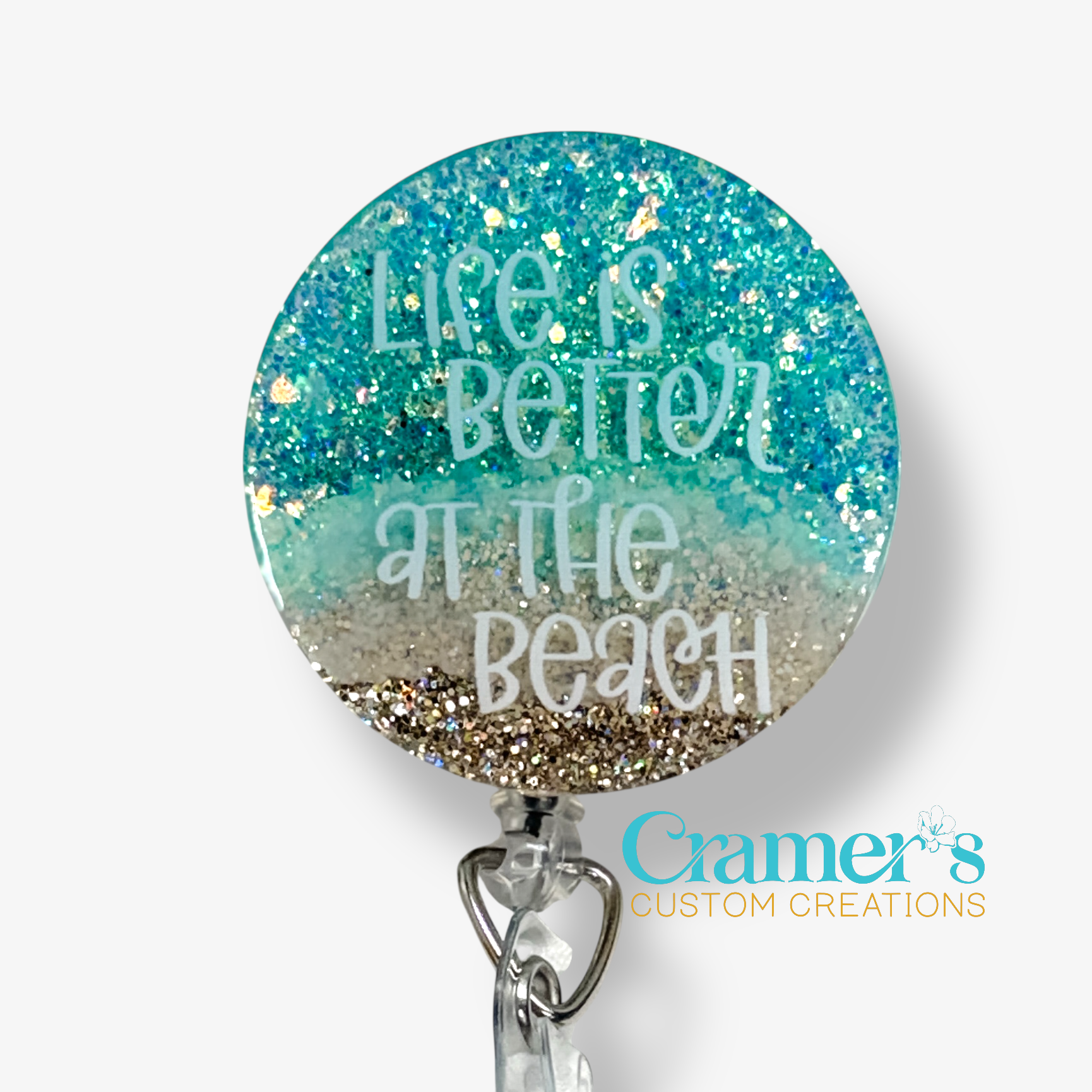 Round badge holder with 'Life is better at the beach' text and glittery design, featuring Cramer's Custom Creations logo.
