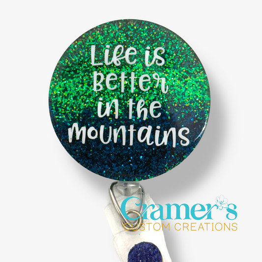 A circle badge reel with a green to blue ombre glitter with the saying life is better in the mountains