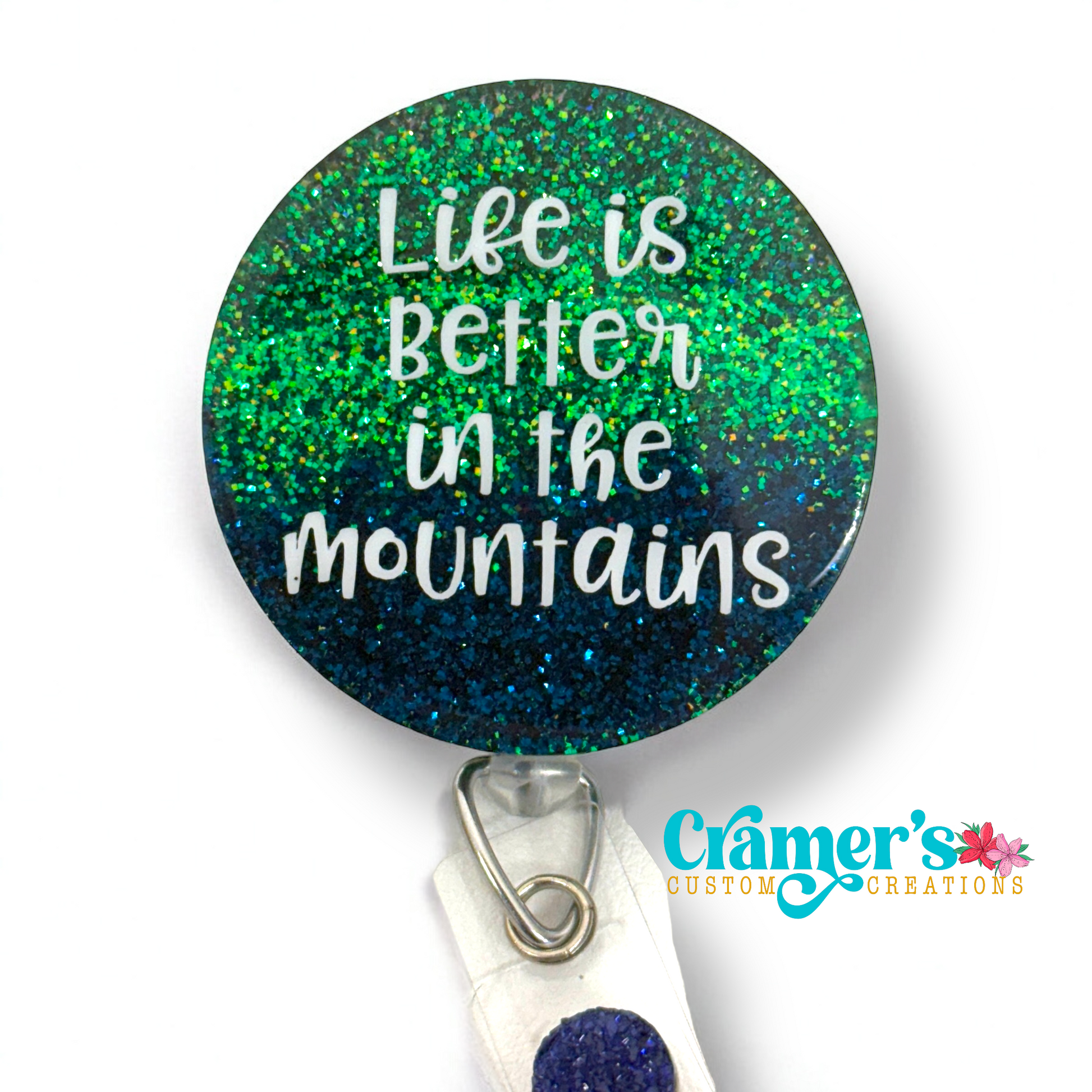 A circle badge reel with a green to blue ombre glitter with the saying life is better in the mountains