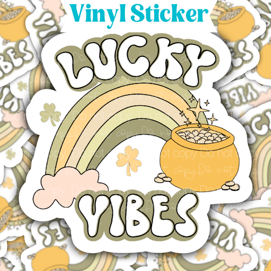 Sticker laying flat that says lucky vibes with a rainbow and pot of gold on a background of the same sticker stacked up