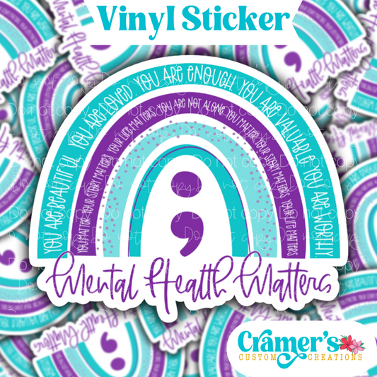 Sticker laying flat that says mental health matter with a rainbow and semi colon in a purple and teal theme, on a background of the same sticker stacked 