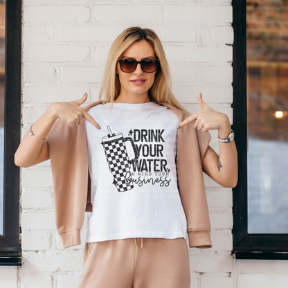 woman wearing a tan sweatsuit standing against a white building pointing to her white shirt that says drink your wanter and mind your business