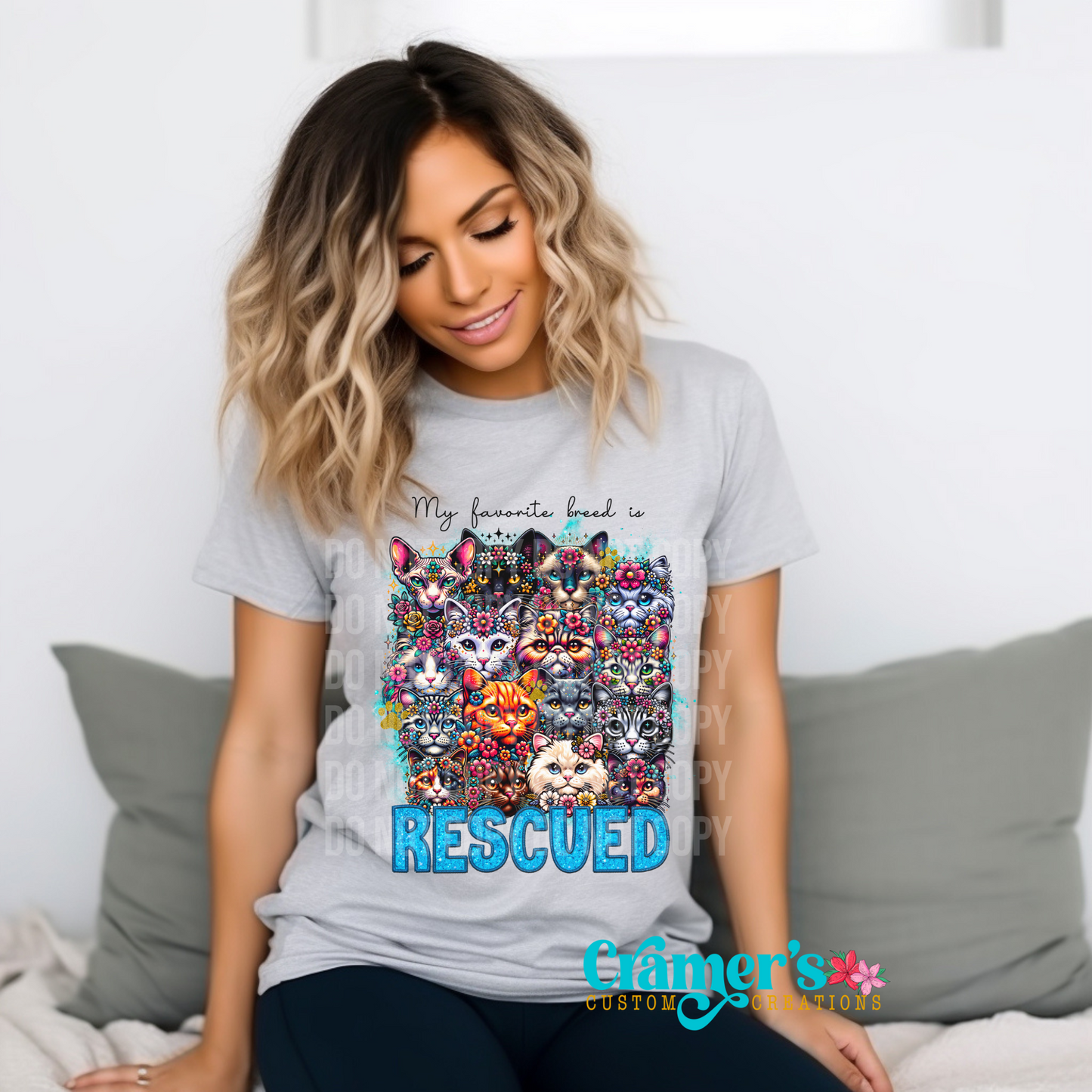 A woman sitting on a couch wearing a gray t-shirt with a colorful graphic print of cats and the text 'MY FAVORITE BREED IS RESCUED (CAT)'.