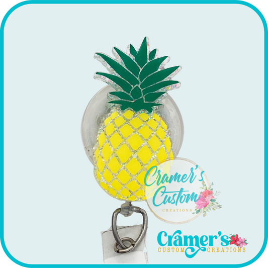 A pineapple-shaped badge reel with a green pineapple top, yellow pineapple body, and a white base, adorned with glitter and alligator clip for attachment.