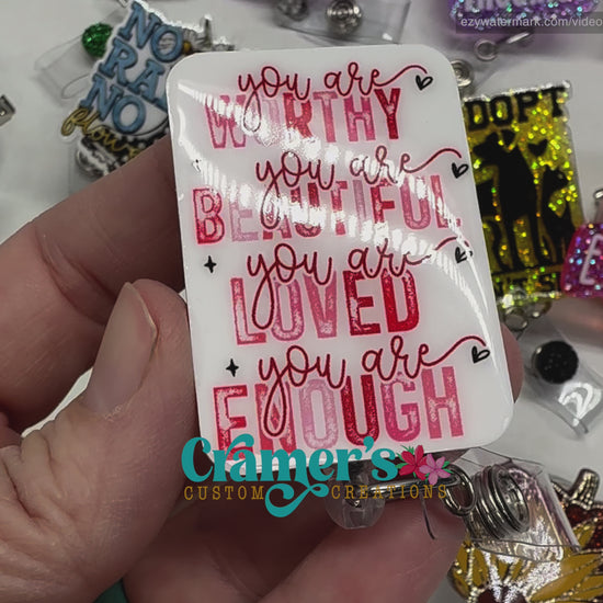 video of a badge reel showing a non glitter option of a badge with the words you are worthy you are beautiful, you are loved, you are enough in pink and red text