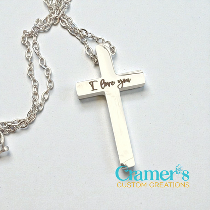 Silver cross necklace with 'I love you' engraving on a light gray background, featuring Cramer's Custom Creations logo.