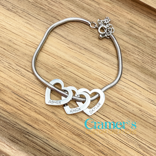 Silver bracelet with interlocking heart charms on a wooden surface, featuring the brand 'Cramer's'.
