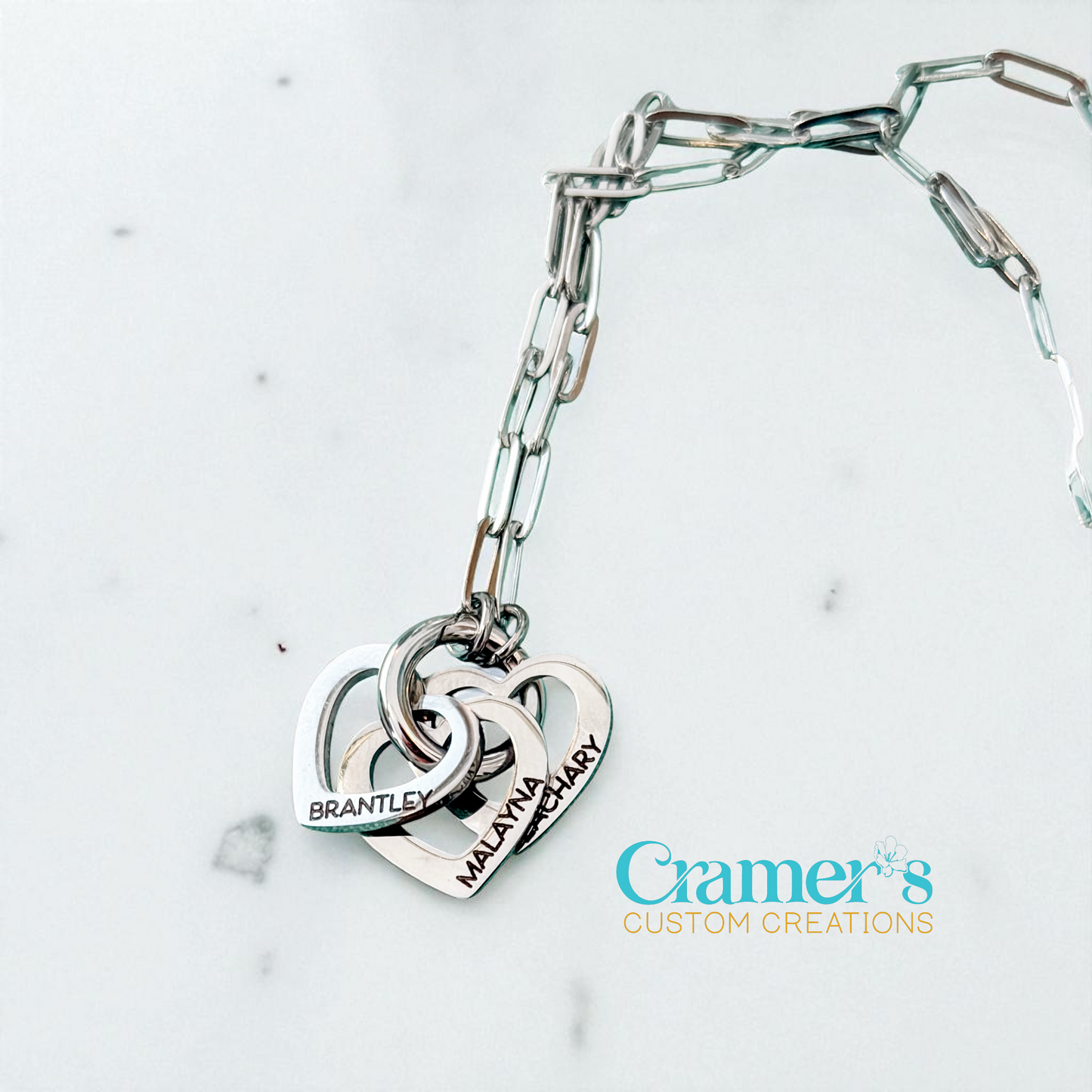 Silver bracelet with interlocking heart charms on a light gray background, featuring 'Cramer's Custom Creations' logo.
