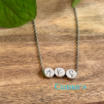 A personalized necklace with the initial 'AKS' on a stainless steel chain with a shiny finish, displayed on a wooden surface with a leaf in the background.