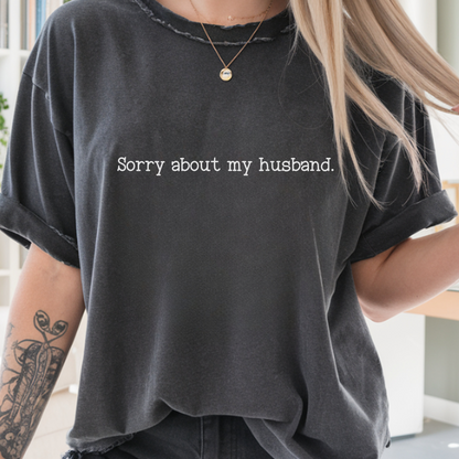A woman wearing a faded black graphic t-shirt with the text 'Sorry about my boyfriend.' printed in white across the chest, paired with black denim shorts in a bright, modern living room setting.