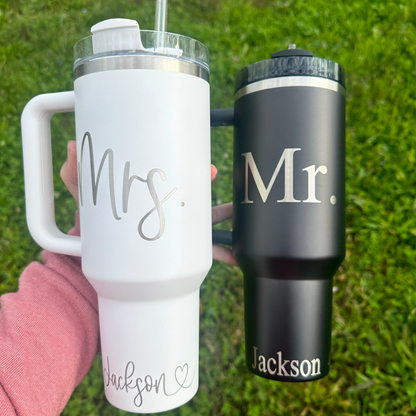 Two engraved tumblers, one white and one black, each with a personalized engraving of 'Mr' and 'Mrs' respectively, held in a person's hand on a grassy background.