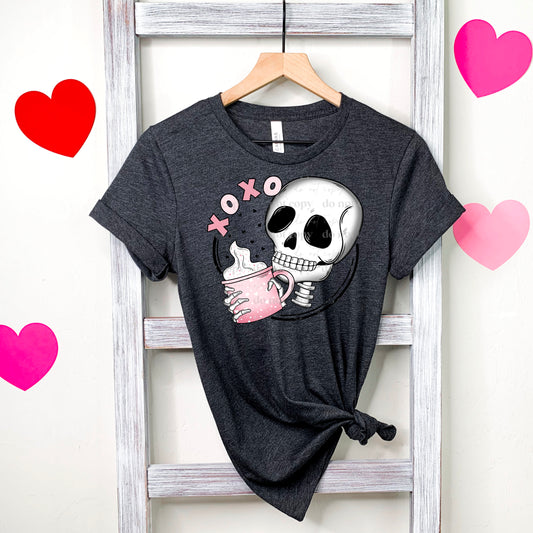 A gray t-shirt with a graphic print of a skull holding a mug, with the text 'XOXO' on the front, displayed on a hanger against a white background with pink heart decorations around.