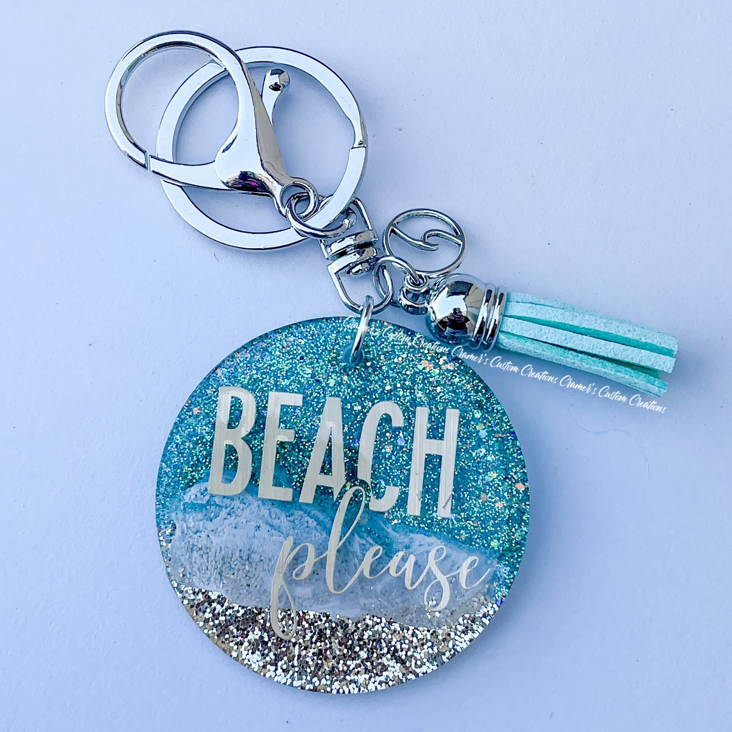 a beachy looking keychain with beach please on it.  It has a wave charm and teal tassel with a lobster claw on a white background