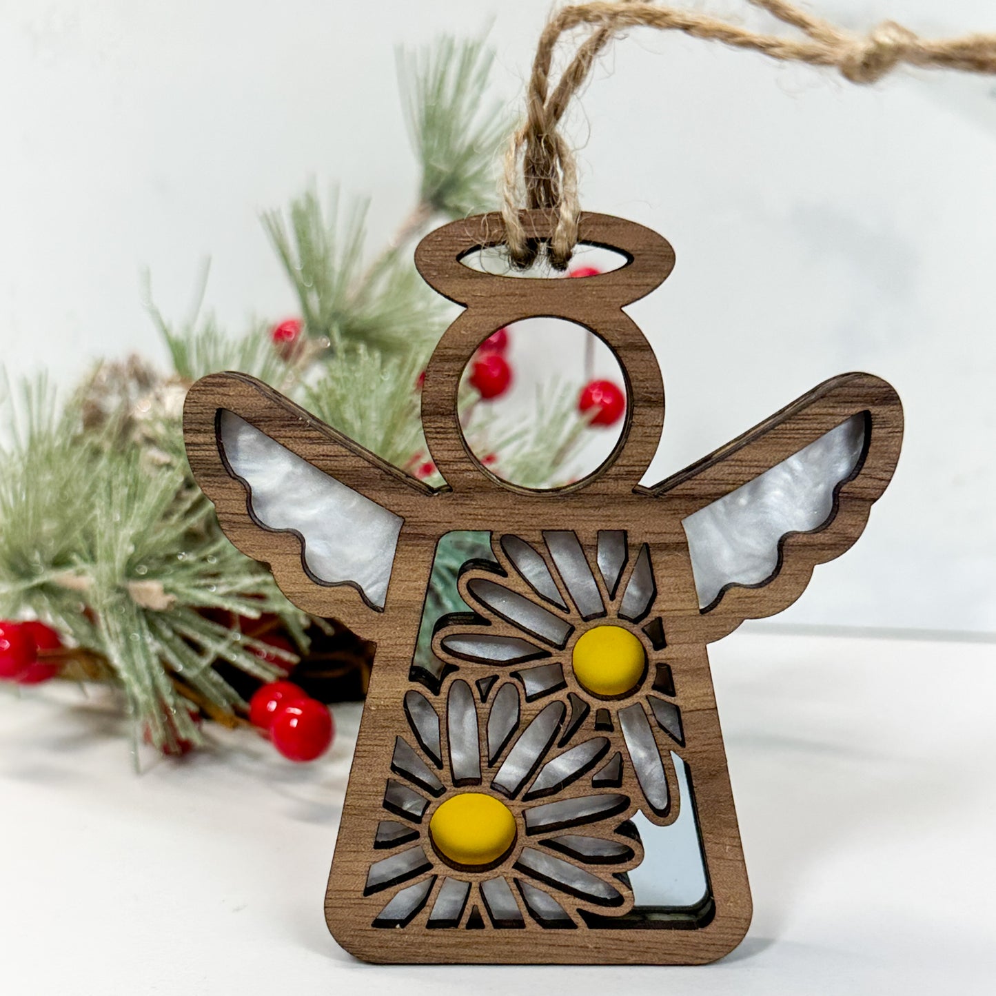 Wooden stained glass looking daisy angel ornament sitting on a white background with pine and berry sprigs