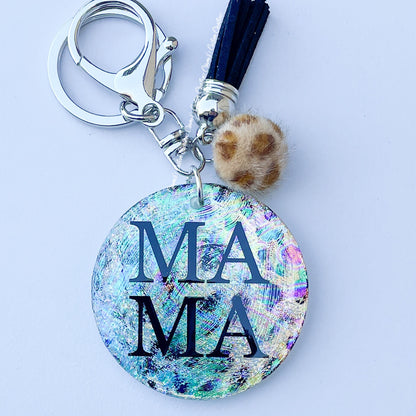 A multicolored glittery keychain with 'MAMA' text and a leopard print design, accented with a tassel and a small jewel.