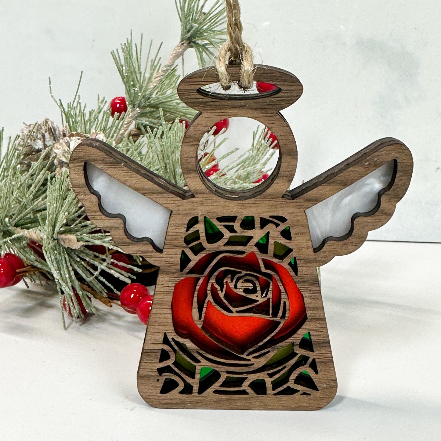 Wooden stained glass looking rose angel ornament sitting on a white background with pine and berry sprigs