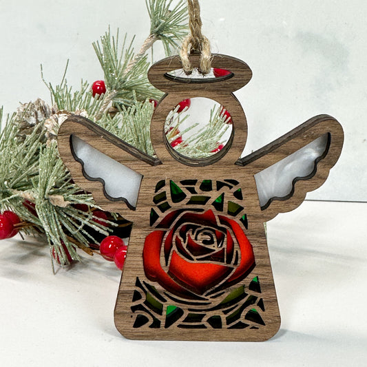 Wooden stained glass looking rose angel ornament sitting on a white background with pine and berry sprigs