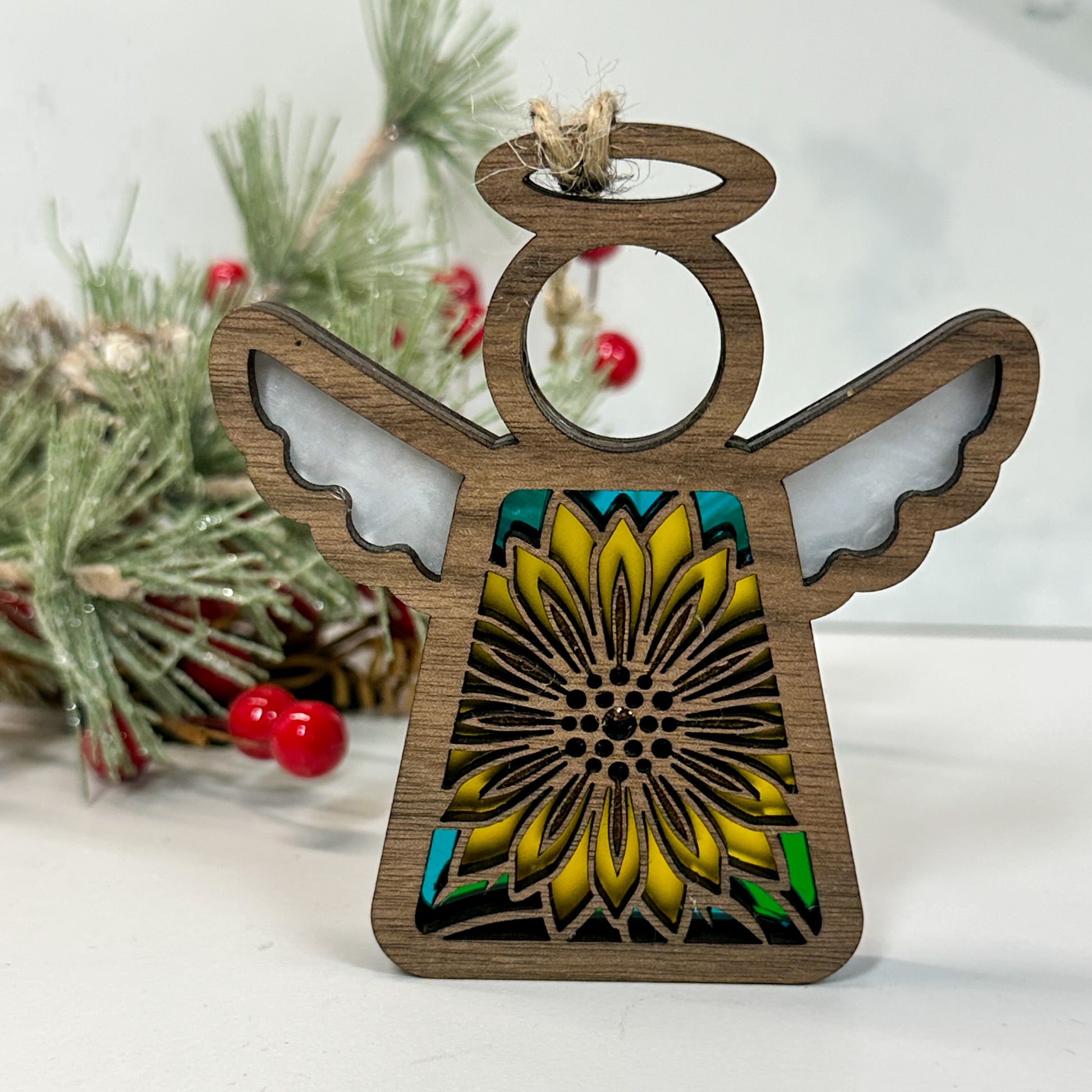Wooden stained glass looking sunflower angel ornament sitting on a white background with pine and berry sprigs