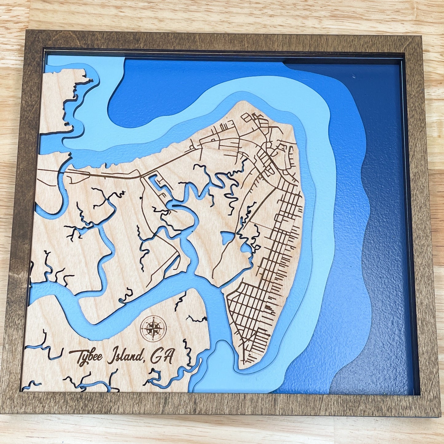 A decorative map framed in a wooden frame, depicting a bathymetric view of Tybee Island, GA, with a blue ocean, brown wood background, and a compass rose at the bottom.
