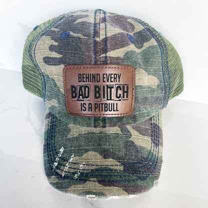 a camo unstructured hat with a rawhide colored patch on the front with behind every bad bitch is a pitbull