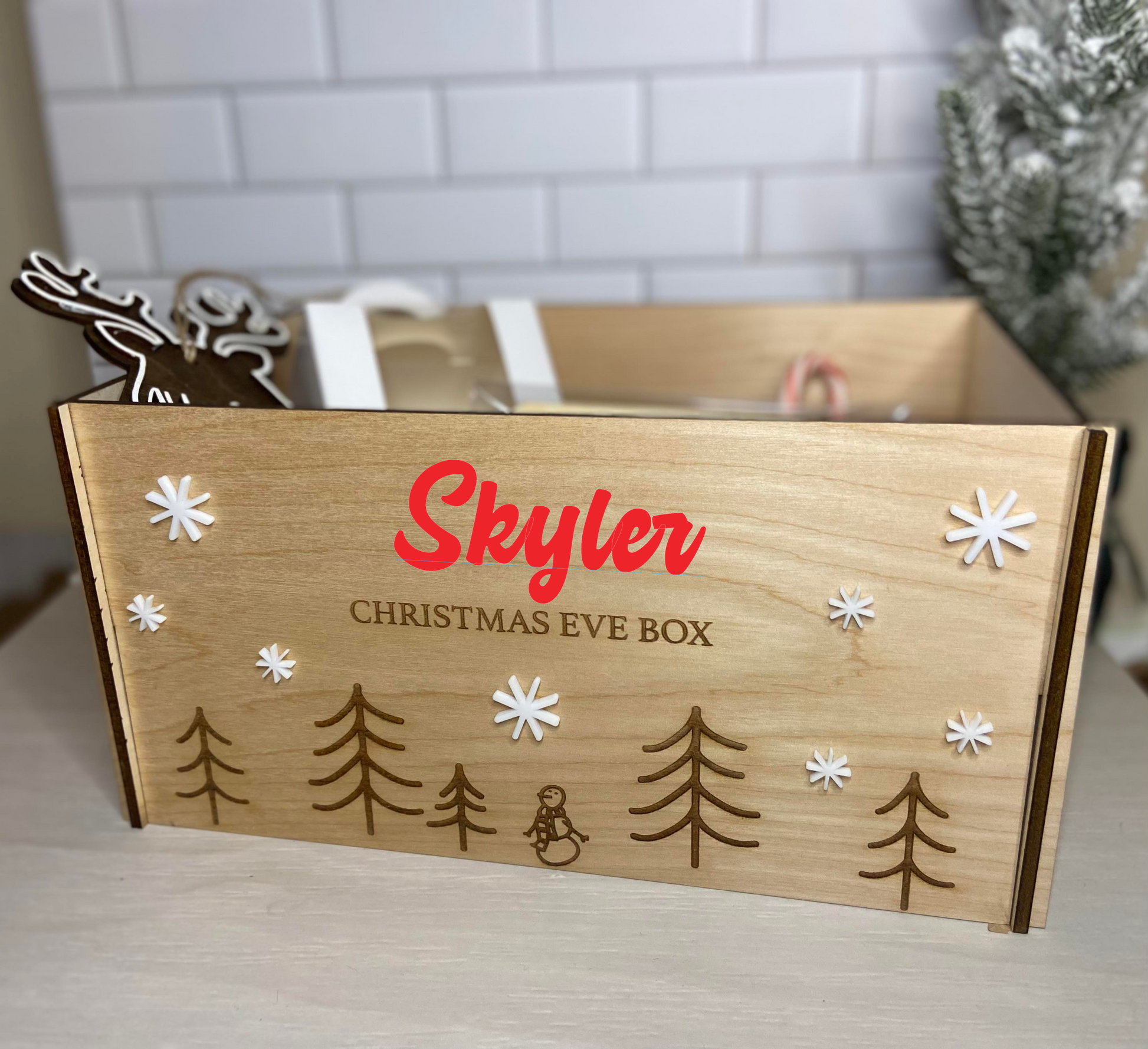 a wood box decorated with snow flakes and engraved trees, and the name skyler on it. on a blurred background
