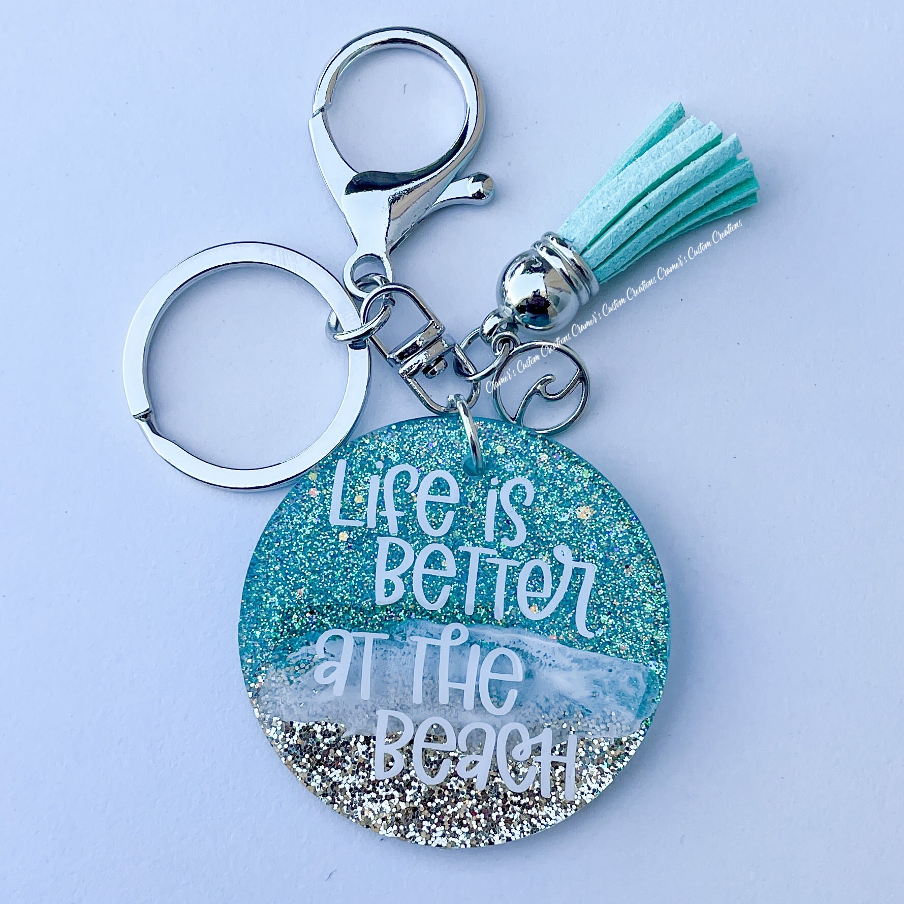 Beachy keychain saying life is better at the beach.  it has a wave charm and a teal tassel with a lobster claw closure on a white background