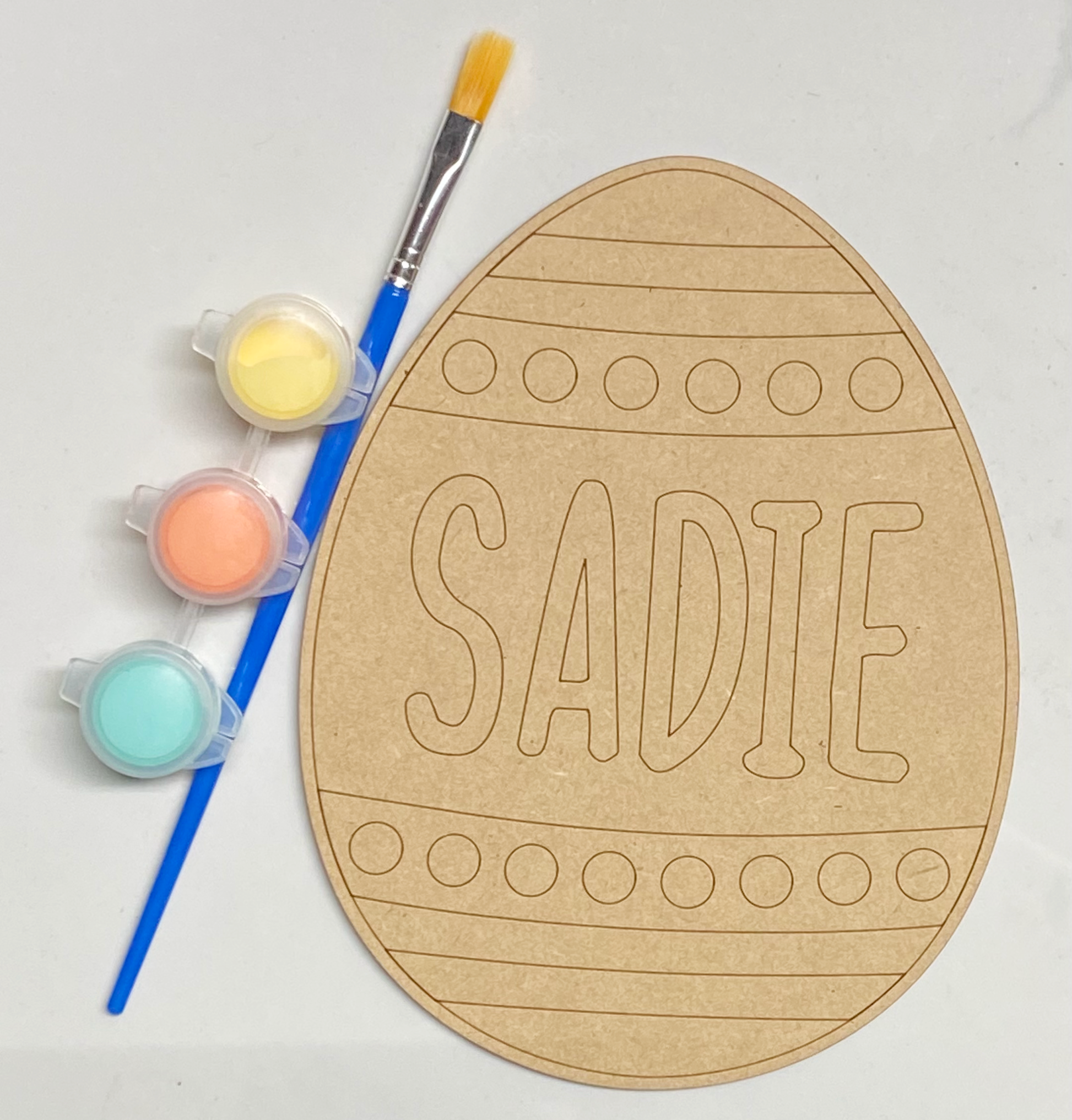 A wooden Easter egg-shaped sign with the name 'Sadie' painted on it, accompanied by three small jars of craft paint and a paintbrush.