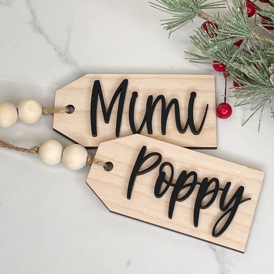 two wood stocking tags with the names mimi and poppy on a white background with pine and berry sprigs