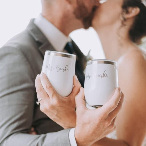 two white wine tumblers with engravings being held up by a bride and groom with them kissing in a blurry background