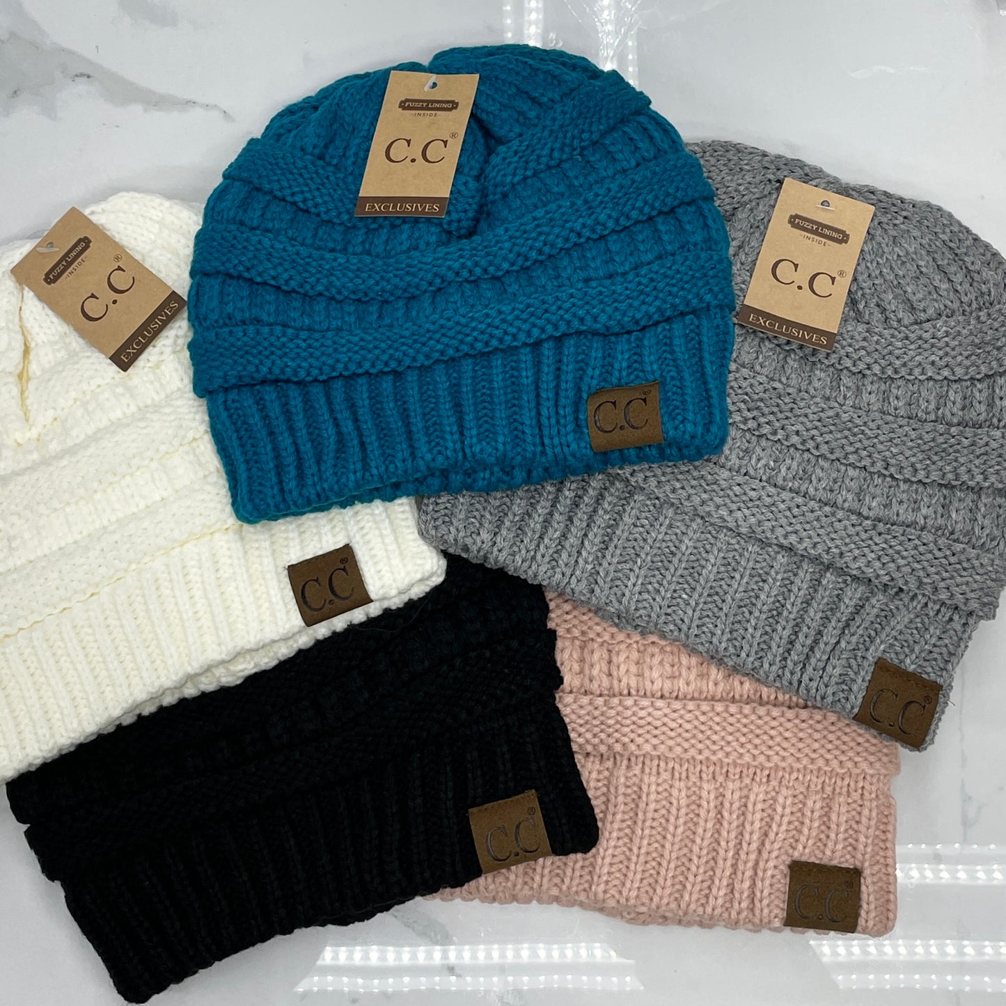 A collection of personalized hats with fuzzy lining, displayed in different colors such as white, blue, gray, black, and pink, each featuring a leather patch with the letter 'CC' on the brim.